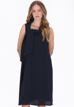 Femeie cu păr lung, ondulat și blond, purtând o rochie midi bleumarin fără mâneci, cu un detaliu de eșarfă legată, pe un fundal alb.