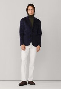 Blazer de pana azul marino, diseño ajustado con dos botones, usado sobre un cuello de tortuga verde oscuro, combinado con pantalones blancos y zapatos marrones.
