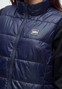 Близък план на човек, носещ тъмносиньо подплатено елече с цип и лого на GAP на гърдите и черна риза с дълъг ръкав отдолу.