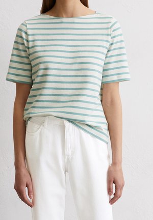 Vrouw draagt een lichtblauw-wit horizontaal gestreept shirt met korte mouwen, ingestopt in een witte broek met hoge taille, tegen een witte muur.