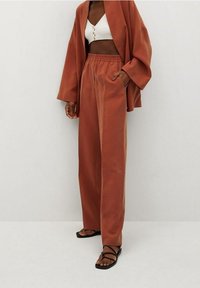 Rdzawoczerwone szerokie spodnie o gładkiej teksturze i elastycznym pasie, w zestawieniu z białą krótką bluzką z guzikami. Czarne sandały na cienkich paskach.