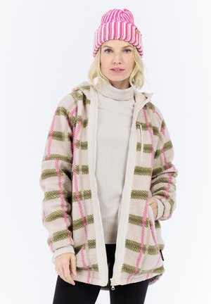 Vrouw met een roze en witte gebreide muts, beige coltrui en een beige, roze en groene geblokte fleece jack, met haar handen in de zakken.