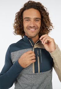 Heren half-zip pullover in marineblauw, beige en grijs. Voorzien van contrasterende panelen, een met rits afsluitbare zak en lange mouwen met een gestructureerde afwerking.