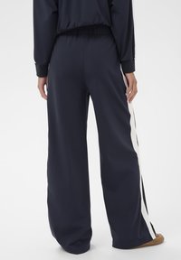 Personne portant un pantalon large noir avec des rayures blanches sur les côtés et un haut noir assorti, se tenant devant un fond blanc uni.