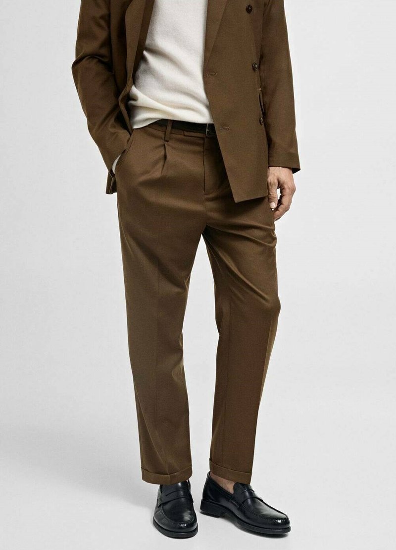 Bruin op maat gemaakt pak, met een dubbelzijdig jasje en geplooide broek, gecombineerd met zwarte leren instapschoenen en een lichte trui.