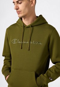 Man in een olijfgroene Champion hoodie met trekkoorden en een voorzak, met zijn handen gedeeltelijk in de zakken, kijkend naar beneden naar de zijkant.