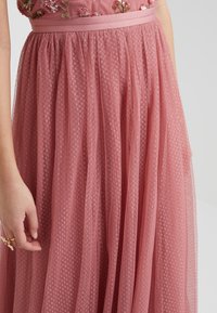 Roze tule jurk met gelaagde, doorschijnende stof. Beschikt over een getailleerde corsage versierd met bloemenapplicaties en een brede tailleband. Textuurpatroon zichtbaar.