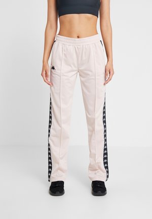 Femme portant un pantalon de survêtement rose clair avec des bandes noires à logo sur les côtés, des chaussures de sport noires et un crop top noir sur un fond uni.