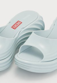 Light blue plastic sandals with an open toe design, layered wave-like sole, and a visible red logo on the insole.