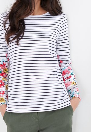 Frau trägt ein weißes Langarmshirt mit schwarzen horizontalen Streifen und Ärmeln mit Blumenmuster, kombiniert mit grünen Hosen.