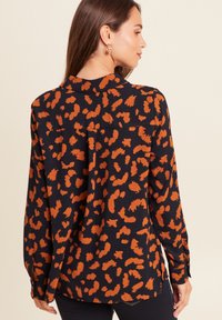 Frau von der Seite dargestellt, die ein langärmeliges schwarzes Shirt mit orangefarbenem Leopardenmuster und goldenen Creolen trägt, vor einem neutralen Hintergrund.