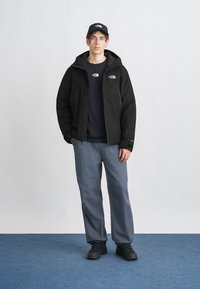 Čierna zateplená bunda s kapucňou, kombinovaná s čiernym tričkom a sivými nohavicami. Celý outfit dopĺňajú čierne tenisky. Minimalistické logo na bunde.