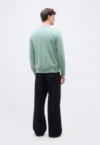 Homme vu de dos, portant un pull à manches longues vert clair et un jean bleu foncé à jambes larges, sur un fond blanc uni.