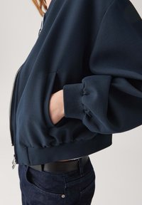 Giacca bomber navy con maniche oversize, polsini a coste e orlo elasticizzato. Presenta una chiusura frontale con zip e tasche laterali per lo stoccaggio.
