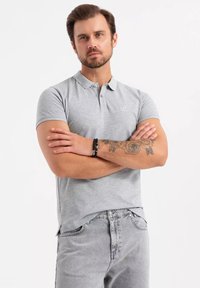 Šedé polo tričko s límečkem, krátkým rukávem a malým logem. Kombinováno s světle šedými džíny, na ruce je viditelné tetování.