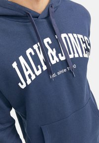 Granatowa bluza z kapturem wykonana z bawełny, z dużym białym nadrukiem "JACK & JONES" na przodzie, z sznurkiem do regulacji i kieszenią z przodu.