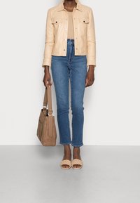 Beige läderjacka med två bröstfickor, ljusbeige figursydd stickad topp, blå medelhöga raka jeans och tanfärgade flätade sandaler.