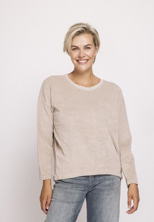 Lächelnde Frau mit kurzen blonden Haaren, die ein beiges Langarmshirt und blaue Jeans trägt, steht vor einem schlichten weißen Hintergrund.
