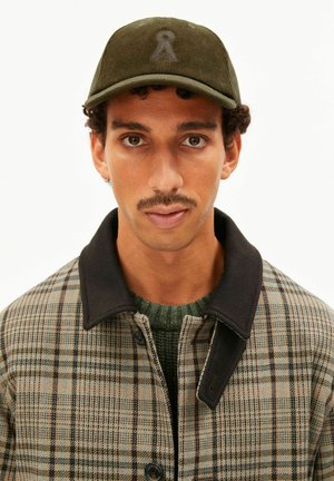 Jonge man met donker krullend haar en snor, die een groene ribfluwelen pet, groene trui en een beige geruite jas met zwarte kraag draagt.