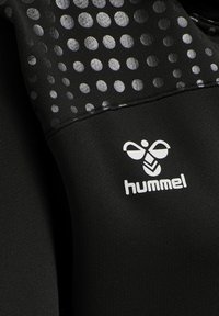 Sort stof med sølvreflekterende polkaprikker og en hvid stiliseret humle-logo over mærkenavnet "hummel".