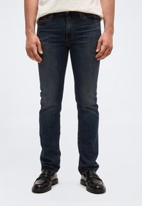 Άνδρας που φοράει στενά τζ ιν σε σκούρο μπλε χρώμα, μαύρα δερμάτινα loafers και λευκό πουκάμισο περασμένο μέσα στο παντελόνι, στέκεται μπροστά σε μια μονόχρωμη επιφάνεια.