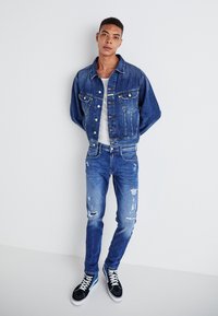Giacca di jeans con vestibilità rilassata e due tasche frontali, abbinata a jeans blu strappati e sneakers nere con lacci bianchi.