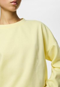 Sudadera amarilla de tela suave con cuello redondo y hombros caídos. Presenta una textura lisa y un ajuste holgado.