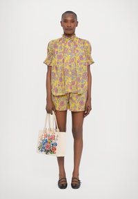 Gul mønstret bluse med korte ermer og matchende shorts, kombinert med en kremfarget håndveske med fargerike broderte blomster.