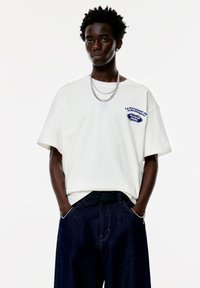 Hvid oversized T-shirt lavet af bomuld, med en blå broderet lap på venstre bryst. Båret med mørke denimbukser og lagde sølvkæder.