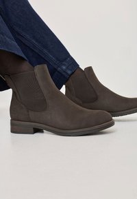 Brown suede ankle boots with elastic side panels, rounded toe, and low block heel. Worn with dark denim jeans, showcasing a casual style.