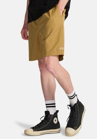 Krótkie spodenki khaki o luźnym kroju z przednimi kieszeniami i małym białym logo. Połączone z czarnymi wysokimi trampkami i paskowanymi skarpetkami.