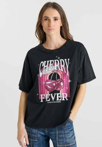 Camiseta negra de gran tamaño con un gráfico de cereza y el texto "FIEBRE DE CEREZA" en blanco y rosa en negrita, con un diseño retro y corte relajado.