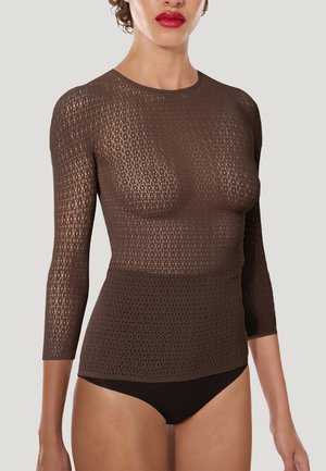 Vrouw die een donkerbruine, lange mouwen, semi-doorzichtige gebreide top met een ronde hals draagt en zwarte ondergoed, tegen een effen lichte achtergrond.