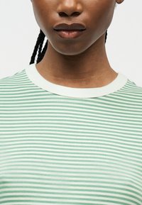 Κοντινό πλάνο ενός ατόμου με πλεξούδες, που φοράει μια λευκή και ανοιχτό πράσινη λεπτοφραγμένη μπλούζα με λευκό γιακά.