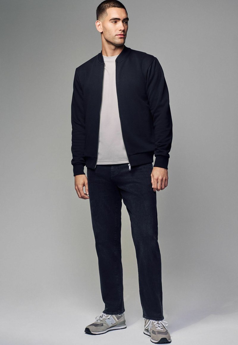 Veste bomber noire sur un t-shirt gris clair, jeans foncés et baskets grises. Look décontracté avec des poignets côtelés et une fermeture éclair à l'avant.