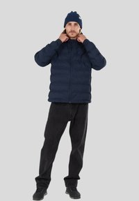 Marineblaue, gepolsterte Jacke mit Reißverschluss, hohem Kragen und seitlichen Taschen, kombiniert mit schwarzen Jeans und einer passenden Strickmütze.