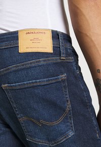 Vaqueros de denim azul con una etiqueta de cuero marrón que dice "JACK & JONES". Las características incluyen costuras naranjas y un bolsillo trasero con una costura curva.