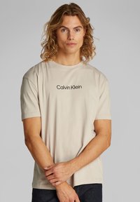 Giovane con lunghi capelli ricci che indossa una maglietta beige di Calvin Klein, in piedi con le mani unite davanti a uno sfondo semplice.