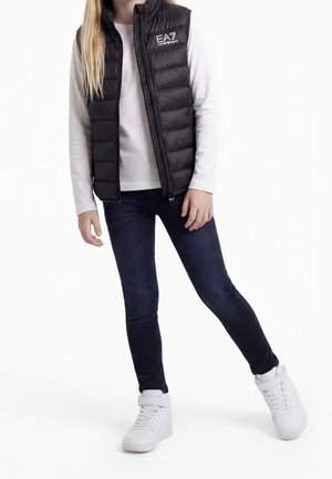 Person iført sort polstret vest med EA7-logo, hvid langærmet skjorte, mørke stramme jeans og hvide høje sneakers.