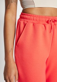 Korallfargede joggebukser med elastisk midje og sidelommer, glatt tekstur og justerbar snøring. Har en tettsittende design ved anklene.