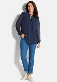 Marineblaue Bluse mit langen Ärmeln, rundem Ausschnitt und teilweisem Knopfverschluss. Kombiniert mit hellblauen Jeans und weißen Sneakers.
