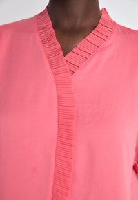 Rosa Bluse mit gefaltetem Ausschnitt, der in V-Form geschnitten ist und aus einem weichen Stoff gefertigt ist. Die Farbe ist lebhaft und einfarbig ohne Muster.