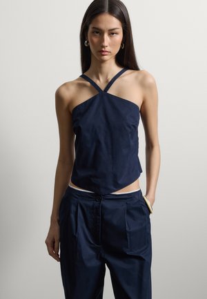 Femme aux longs cheveux bruns portant un haut sans manches bleu marine à bretelles croisées et un pantalon taille haute assorti sur un fond uni.