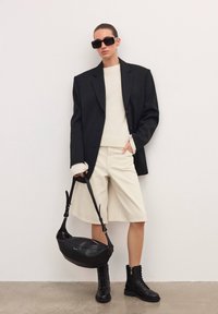 Czarna, oversize'owa marynarka, jasny kremowy sweter, beżowe szorty, czarne wojskowe buty oraz czarna torba na ramię z zamkiem. Tło jest jednolicie białe.