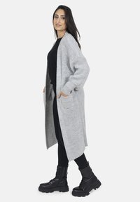 Grijs gebreide lange cardigan met open voorkant, zijzakken en doorhangende schouders, gecombineerd met een zwarte top en legging, aangevuld met zwarte laarzen.
