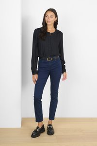 Černá dlouhá košile na knoflíky, tmavě modré skinny džíny, černé polobotky se zlatými akcenty, stojící na světlé dřevěné podlaze u bílé zdi.