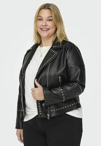 Svart fusk läder bikerjacka med silvernitar på axlarna och ärmarna, fickor med dragkedja och en figurnära design. Bärs över en vit skjorta.