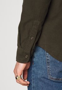 Olivgrön manchester-skjorta med knäppta manschetter, sedd från sidan, kombinerad med blå denimjeans. Guldring på handen med detaljerad struktur.