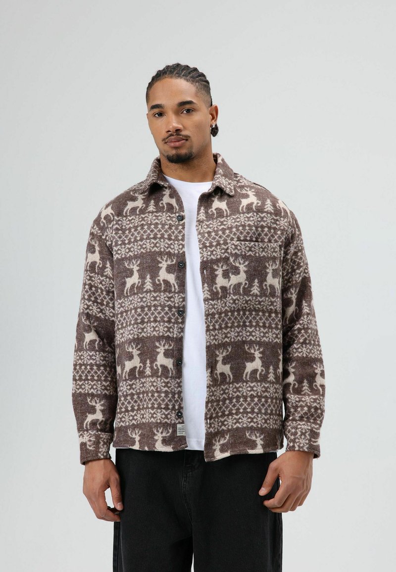 Veste-chemise marron à motifs avec des cerfs crème et des arbres, boutonnée à l'avant, avec col et une poche poitrine. Portée sur une chemise blanche.