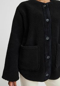 Svart fleecejacka med strukturerad yta, med en mörk kordfläktkläpp, fem knappar och två framfickor. Mjuk, oversized passform.